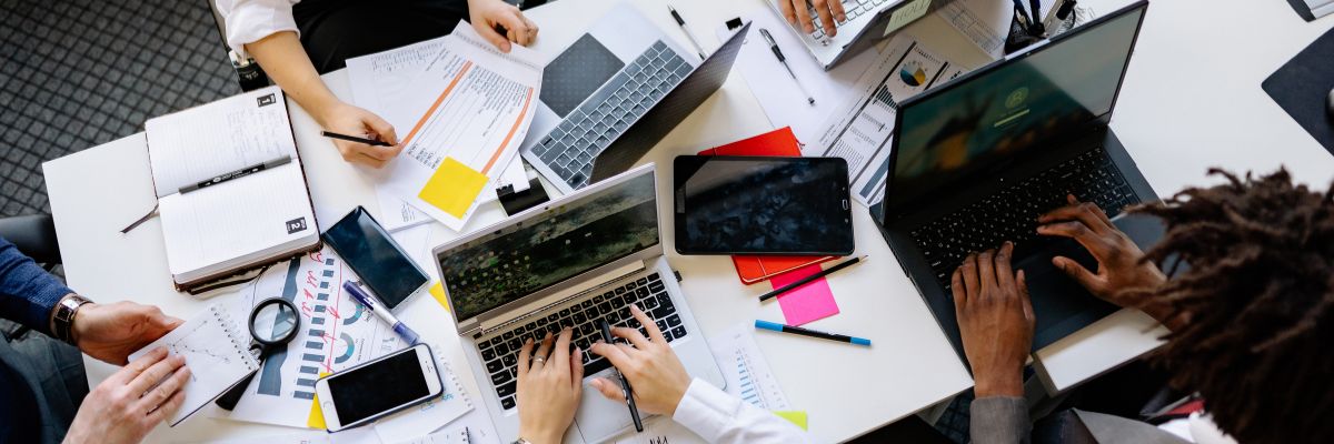 A top-down view of a busy white desk with multiple people using laptops, tablets, and reviewing printed reports.