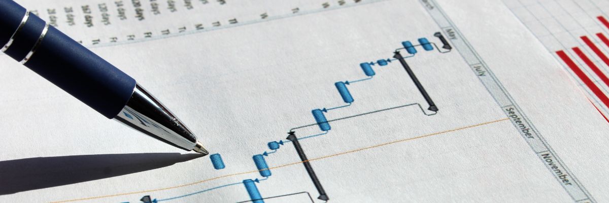 A close-up of a blue pen pointing to a specific task on a printed Gantt chart or project schedule.