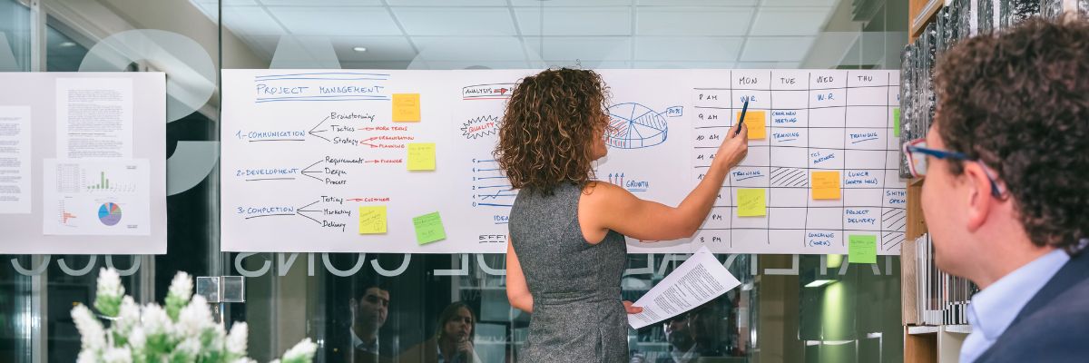 A woman standing at a large whiteboard covered in project management diagrams, schedules, and colorful sticky notes.