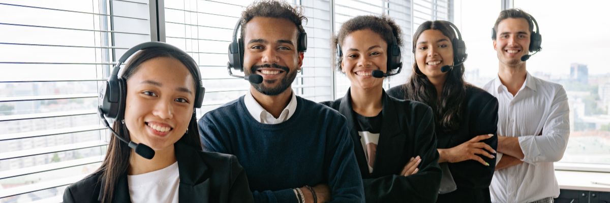A diverse group of five smiling professionals wearing headsets, standing together in a bright office.