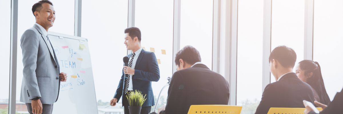 A man speaking into a microphone while standing next to a whiteboard during a team training session.